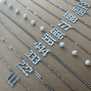 以下が含まれることがあります： 真珠のアクセントが付いた銀のネックレス9点セット。各ネックレスには、ゴシック体で異なる数字が刻まれています。数字は11、22、33、44、55、66、77、88、99です。