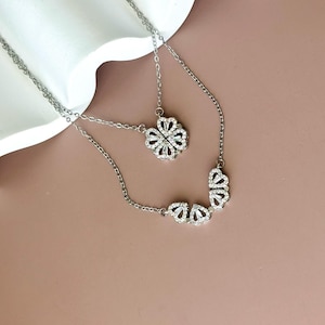 K&ouml;nnte beinhalten: Eine silberfarbene Halskette mit einem Anh&auml;nger, der sich zwischen einem vierbl&auml;ttrigen Kleeblatt und einer Reihe aus vier Herzen verwandeln l&auml;sst. Die Metallkette und die herzf&ouml;rmigen Segmente sind mit kleinen, klaren Steinen besetzt.