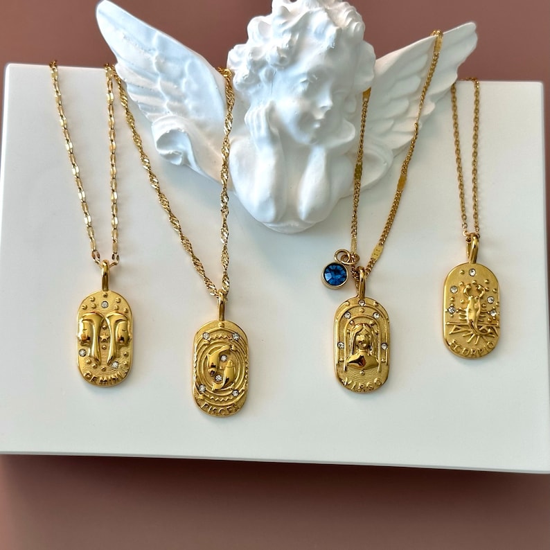 May include: Four gold-toned necklaces with oval pendants featuring zodiac signs Gemini, Pisces, Virgo, and Scorpio. Each pendant is embossed with a symbol, small clear crystals, and the name of the sign. One necklace includes a small blue crystal charm.