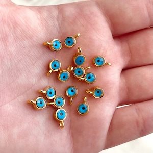 May include: A collection of gold-toned evil eye charms with blue centers. The charms are scattered on a hand.