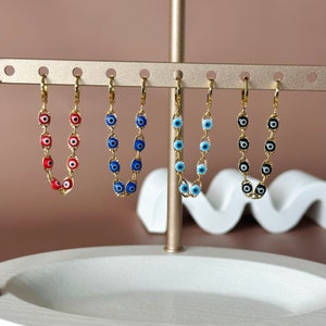May include: Five gold chain earrings with blue and white evil eye charms. The charms are in different colors: red, blue, light blue, black, and white.