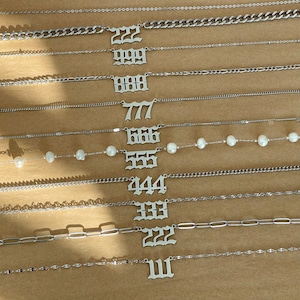 以下が含まれることがあります： 111、222、333、444、555、666、777、888、999、22 などの数字が刻まれたシルバーネックレスのセット。一部のネックレスはチェーンリンクのデザイン、他のネックレスはより繊細なチェーン。1つのネックレスには白いパールが飾られています。