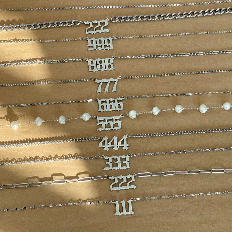 以下が含まれることがあります： 111、222、333、444、555、666、777、888、999、22 などの数字が刻まれたシルバーネックレスのセット。一部のネックレスはチェーンリンクのデザイン、他のネックレスはより繊細なチェーン。1つのネックレスには白いパールが飾られています。