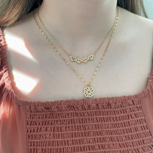 K&ouml;nnte beinhalten: Ein goldfarbenes, mehrlagiges Halskettenset mit zwei zarten Ketten. Die k&uuml;rzere Kette zeigt vier kleine, herzf&ouml;rmige Anh&auml;nger, die mit klaren Steinen besetzt sind. Die l&auml;ngere Kette hat einen kleeblattf&ouml;rmigen Anh&auml;nger mit klaren Steinen.