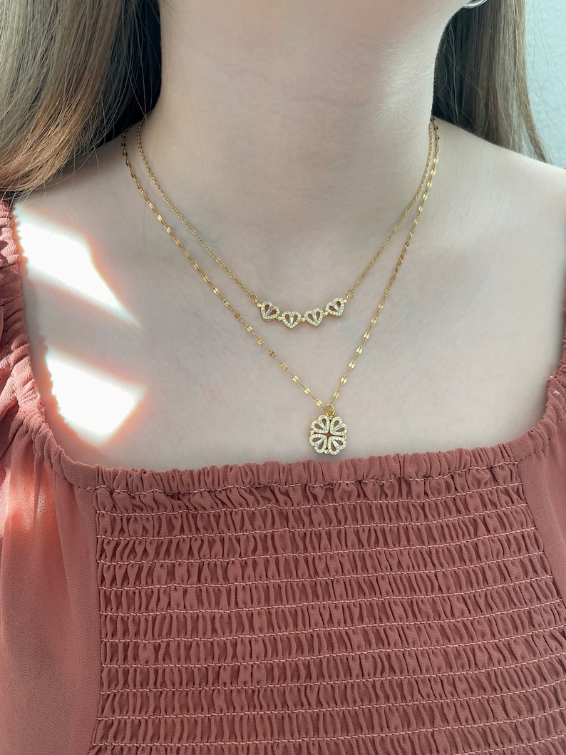 K&ouml;nnte beinhalten: Ein goldfarbenes, mehrlagiges Halskettenset mit zwei zarten Ketten. Die k&uuml;rzere Kette zeigt vier kleine, herzf&ouml;rmige Anh&auml;nger, die mit klaren Steinen besetzt sind. Die l&auml;ngere Kette hat einen kleeblattf&ouml;rmigen Anh&auml;nger mit klaren Steinen.