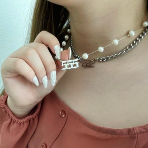 以下が含まれることがあります： シルバーチェーンネックレスに、"555"と書かれたシルバーペンダントと、"222"と書かれた小さなペンダントが付いています。ネックレスはパールネックレスの上に重ねられています。