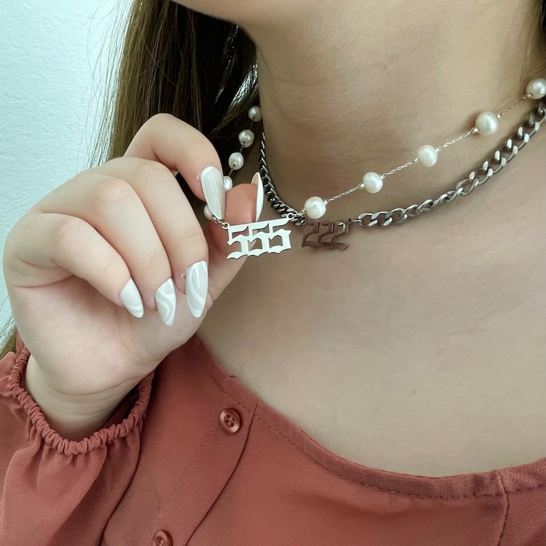 以下が含まれることがあります： シルバーチェーンネックレスに、"555"と書かれたシルバーペンダントと、"222"と書かれた小さなペンダントが付いています。ネックレスはパールネックレスの上に重ねられています。