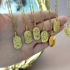 May include: A collection of gold-toned necklaces featuring oval-shaped pendants with embossed zodiac symbols and small embedded clear crystals. Each pendant is suspended from a twisted gold chain, displaying signs including Aquarius, Pisces, Aries, and Taurus.