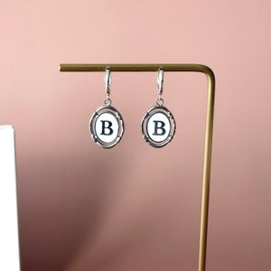 May include: A pair of silver hoop earrings with a white oval charm featuring the letter "B" in black.