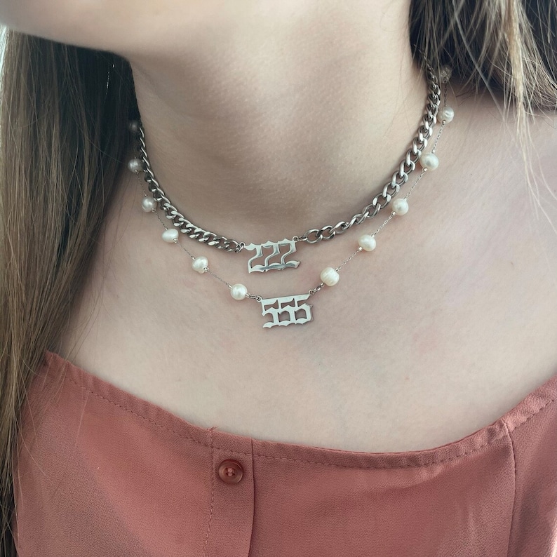 May include: A layered necklace set featuring a silver-toned curb chain with a pendant displaying the number 222 in an old English font, and a delicate chain adorned with small white pearls and a pendant displaying the number 555 in old English.