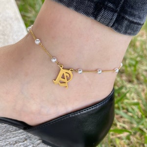 May include: Gold-toned anklet with small white pearls and a gold letter charm. The anklet is worn on an ankle, paired with a black shoe and denim jeans. The charm features the letters "P" and "A".