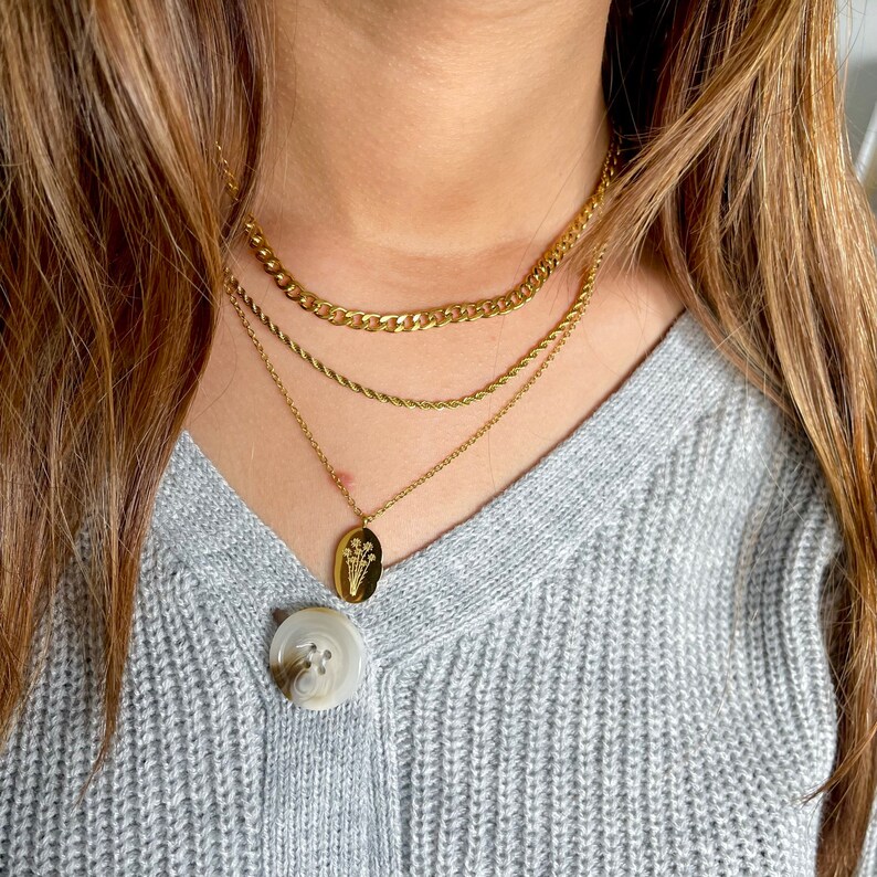 May include: Gold layered necklaces. The top layer is a curb chain, the middle is a rope chain, and the bottom features a pendant with a floral design. The necklaces are worn over a grey sweater.