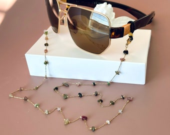 May include: Gold-rimmed sunglasses with brown gradient lenses and dark tortoiseshell temples, attached to a delicate gold-tone chain featuring small, multi-colored natural stone beads in shades of green, pink, and black, draped elegantly.