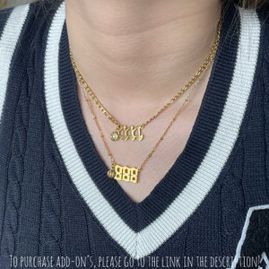 May include: Two gold-toned layered necklaces featuring gothic-style number pendants. The top necklace displays the number 444 with a small green circular charm, while the bottom necklace features the number 888 with a small circular Pisces symbol charm.