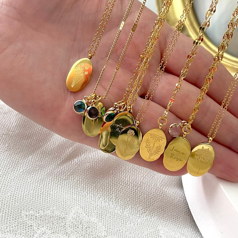 May include: A selection of gold-coloured necklaces, each with an oval pendant. The pendants display floral designs and hang from fine chains. Some necklaces have gemstone embellishments. The words "June Rose" are engraved on one pendant.