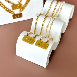 May include: Three gold necklaces with different pendant styles. The pendants are rectangular with a textured edge and feature small gemstones. The necklaces are displayed on a white wavy surface.
