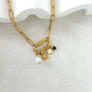 Op de afbeelding: Goudkleurige ketting met een grote sluiting en schakels. De ketting heeft een parel, een gouden letter "K", een hartbedel en een blauwe oogbedel. De ketting wordt gepresenteerd op een witte ondergrond.