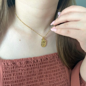 May include: A gold-toned necklace featuring a thin, twisted chain and a small, rectangular pendant. The pendant displays a raised, embossed design of a ram's head, representing the Aries zodiac sign, accented with small, clear, sparkling stones.