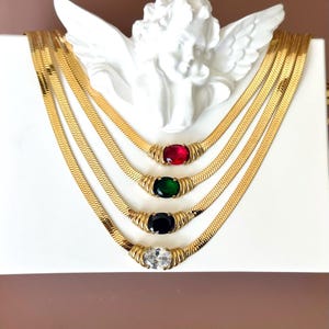 May include: Four gold-colored herringbone necklaces with oval-shaped gemstones in red, green, black, and clear. The necklaces are displayed on a white surface with a decorative angel statue in the background.