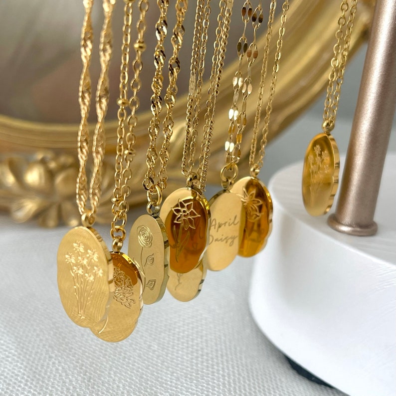 May include: A selection of gold-coloured necklaces, each with an oval pendant. The pendants display floral designs and hang from fine chains. Some pendants are engraved with the words "April Daisy".