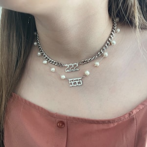 以下が含まれることがあります： 人の首に重ねられた2本の銀のネックレス。上のネックレスは太い銀のチェーンで、「222」と書かれたペンダントが付いています。下のネックレスは繊細なチェーンで、小さな白い真珠と「555」と書かれたペンダントが付いています。