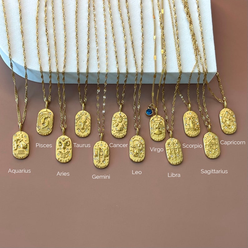 May include: A collection of gold-toned necklaces featuring oval pendants, each embossed with a unique zodiac sign and small embedded crystals. The pendants are labeled with the names Aquarius, Pisces, Aries, Taurus, Gemini, Cancer, Leo, Virgo, Libra, Scorpio, Sagittarius, and Capricorn.