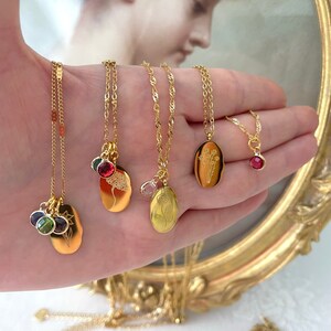 May include: Several gold necklaces with different pendant styles. The pendants feature a sun, a tree, a flower, and a bouquet, some with colourful gemstones. The necklaces are shown on a hand.