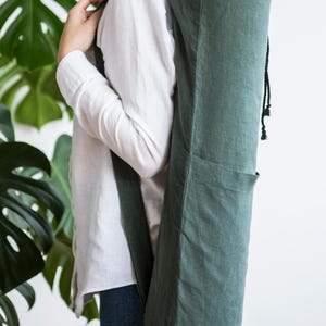 Op de afbeelding: Een donkergroene yogamat-tas van linnen. De tas heeft een trekkoordsluiting aan de bovenkant en een zak aan de zijkant. De tas wordt over de schouder gedragen. Ongeveer 80 cm lang.