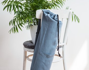 May include: A blue yoga mat bag with a drawstring closure hangs on the back of a white chair. A potted plant with green leaves sits behind the chair. The bag is made of a soft, textured fabric.
