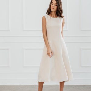 May include: A woman wearing a sleeveless, off-white midi dress with a simple, rounded neckline. She is standing on a white tiled floor and wearing silver heeled sandals.