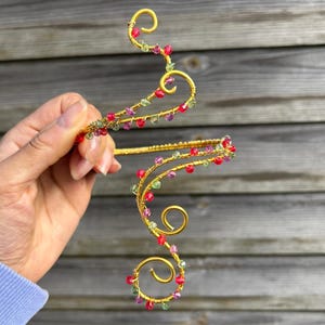May include: A gold-colored wire headpiece with a swirling design, adorned with red, green, and purple beads. The headpiece is held up against a wooden background, showcasing its intricate, handcrafted details. The design features a series of loops and curves.
