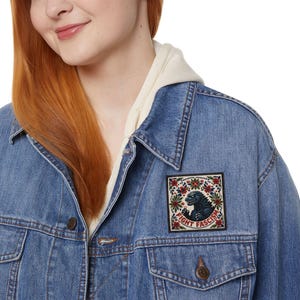 May include: A light blue denim jacket with a patch featuring a Godzilla-like creature and the words "FIGHT FASCISM." The patch is surrounded by a floral design in red and green. The jacket has a button-down front and two chest pockets.