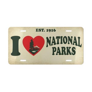Puede incluir: Placa de matrícula con un diseño vintage. La placa presenta el texto "I ❤️ NATIONAL PARKS" en verde oscuro y blanco, con un corazón rojo que contiene una ilustración de montaña y árbol. El texto "EST. 1916" está en la parte superior.