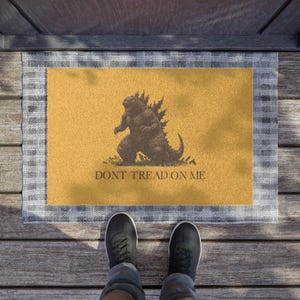 Puede incluir: Una alfombra rectangular con un gráfico de Godzilla sobre un fondo amarillo, enmarcada por un borde a cuadros gris y blanco. El texto "DONT TREAD ON ME" está debajo. Se ven zapatos negros.