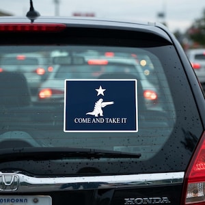 Puede incluir: Una pegatina rectangular en la ventana trasera de un coche. La pegatina tiene un fondo azul oscuro con una estrella blanca y una criatura parecida a Godzilla sosteniendo un rifle. El texto "COME AND TAKE IT" está debajo.