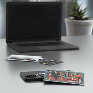 May include: A black smartphone with a silver ring holder, a small rectangular keychain with a Japanese art print, a white card holder, and a black laptop on a gray surface. A credit card and a small potted plant are also visible.