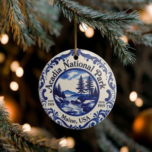 May include: A round white ceramic ornament featuring a blue illustration of Acadia National Park in Maine, USA. The text reads Acadia National Park, ESTD 1919, and Maine, USA. It has a blue decorative border and hangs from a thin gold string.