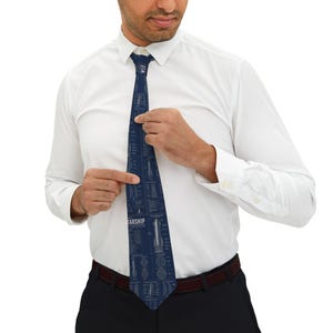 Peut inclure: Une cravate bleu foncé ornée d'un motif de plan technique blanc représentant une fusée. Le design comprend des schémas détaillés, des étiquettes techniques et le mot STARSHIP imprimé en blanc. La cravate est portée sur une chemise blanche.
