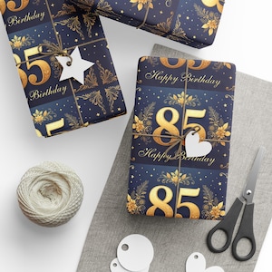 Könnte beinhalten: Geschenke, verpackt in marineblauem Papier mit goldenen Blumenmustern und der Zahl 85. Die Geschenke sind mit Bindfaden gebunden und mit weißen herz- und sternförmigen Anhängern versehen. Eine Garnrolle und eine Schere sind ebenfalls zu sehen.