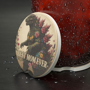May include: A round car coaster featuring a floral Godzilla design and the text "Best Mom Ever." The coaster has a cream border and is next to a red container with water droplets.