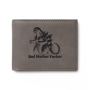 May include: Gray leather wallet with a stitched border. The wallet features a black illustration of Godzilla and the text "Bad Mother Fucker" in a bold font.