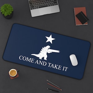 Puede incluir: Alfombrilla de escritorio rectangular azul marino con un gráfico blanco de Godzilla sosteniendo un cañón, una estrella blanca y el texto "COME AND TAKE IT". Un ratón de ordenador blanco está sobre la alfombrilla.