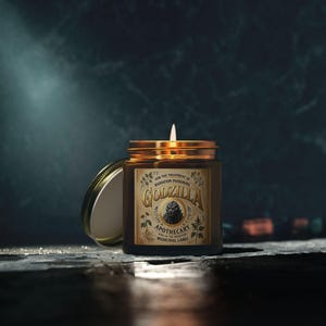 May include: A brown glass jar candle with a gold lid and a lit white wick. The label reads "Godzilla Apothecary" with a monster illustration and text "For the treatment of radiation poisoning". The lid is partially open, revealing the candle's interior.