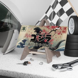 May include: A decorative license plate with a Godzilla design, Japanese text, and cherry blossoms. The plate is displayed on a wooden stand, with a checkered flag and a tyre in the background. A helmet, spanner, and keys are also visible.