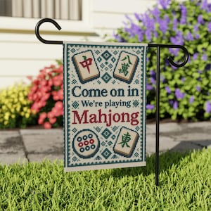 Puede incluir: Una bandera de jardín rectangular con un patrón de punto de cruz y bordes geométricos azules y verdes. El texto Come on in We’re playing Mahjong aparece en azul y rojo. Cuatro fichas de Mahjong se muestran en las esquinas del diseño.