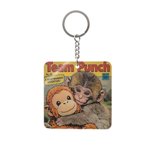 May include: Square keychain featuring a comic book cover with two monkeys hugging. The text "Team Punch" is at the top, with "No. 24" and "A Heartwarming Adventure!" below. A silver chain and keyring are attached.
