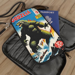 Puede incluir: Una cartera de viaje negra con una ilustración de Godzilla en la parte delantera. La ilustración es de un póster japonés vintage. La cartera tiene una cremallera y una correa.