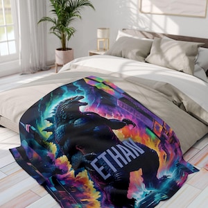 May include: A black and blue throw blanket with a Godzilla-like monster in a colorful, neon-like background. The name "ETHAN" is written in white letters on the blanket.