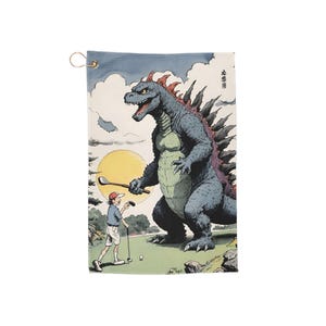 May include: Golf towel with a cartoon illustration of a Godzilla-like monster playing golf. The monster holds a golf club, with a golfer in the background. The design is set against a sunny sky and green golf course. A gold-coloured metal ring is attached.