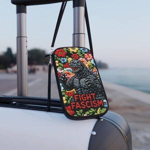 May include: Black crossbody bag featuring a Godzilla graphic surrounded by colorful flowers. The bag has the text "FIGHT FASCISM" in red and white. The bag is hanging from a black strap.